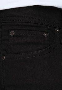Vaqueros de mezclilla negra con una textura suave, diseño clásico, herrajes en tono cobre y costuras sutiles en los bolsillos.
