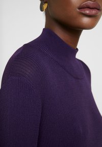 Gros plan d'une personne portant un pull en maille violet côtelé avec un col montant et une boucle d'oreille circulaire dorée.