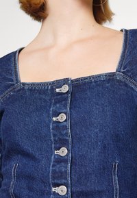 Gros plan sur un haut en denim avec des boutons argentés sur le devant et un col carré porté par une personne à la peau claire et aux cheveux courts.