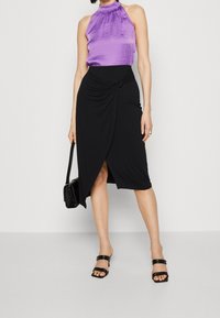 Top halter in raso viola abbinato a una gonna avvolgente nera con un dettaglio a nodo. Indossato con sandali neri con tacco a blocco e una piccola borsa.