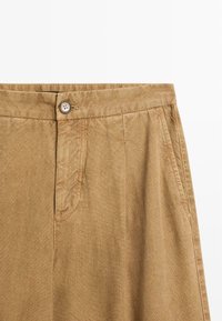 Close-up of tan cotton trousers showing waistband with button, front zipper, and side pockets on a white background.