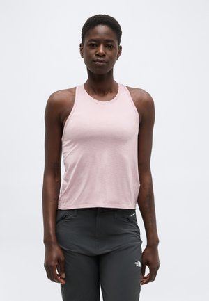 Femeie purtând un top roz pal fără mâneci și pantaloni gri închis North Face, stând în picioare pe un fundal simplu, deschis.