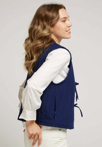 Gilet matelassé navy avec liens sur les côtés, doté d'un col rond et de détails de couture complexes. Porté par-dessus une blouse blanche et un pantalon clair.
