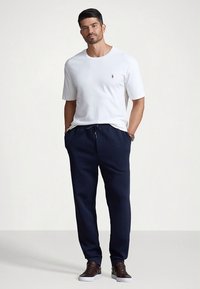 Balti trumpomis rankovėmis marškinėliai su mažu logotipu, derinami su tamsiai mėlynomis sportinėmis kelnėmis, kurių liemuo elastingas, o šonuose yra kišenės. Rudos sportbačiai.