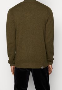 Olivgrön stickad tröja med ribbat halsringning och ärmslut. Har en märkesetikett vid den nedre fållen och en avslappnad passform.