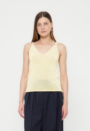 Žena s ravnom smeđom kosom, koja nosi zlatne naušnice u obliku obruča, blijedo žuti pamučni top bez rukava i tamne plisirane hlače, stoji ispred bijele pozadine.