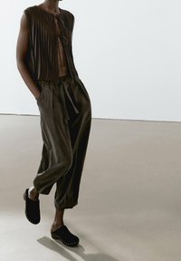 Brown pleated sleeveless top with a tie closure, paired with loose dark trousers and black clogs featuring metal studs along the edge.