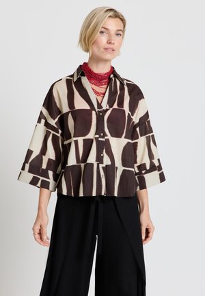 Femme blonde portant une chemise à motifs noirs et beiges à boutons surdimensionnés, un pantalon noir à jambes larges et un collier superposé de perles rouges.