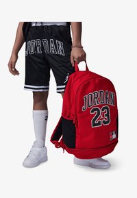 Sac à dos rouge avec des accents noirs, arborant "JORDAN 23" en lettres grasses. Il comprend une texture lisse et des bretelles ajustables, tenu à la main.