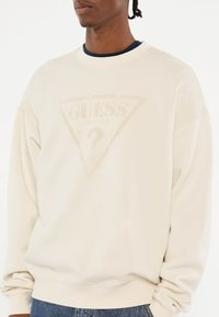 Beige crewneck sweatshirt met een vervaagde driehoekige logo in het midden, gemaakt van zacht materiaal met ribgebreide manchetten en zoom.