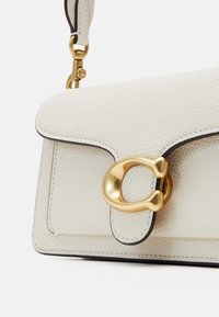 Sac à main compact en cuir blanc cassé avec texture granulée, bordure noire et fermoir en métal doré sur le rabat avant, attaché à une bandoulière avec un crochet doré.