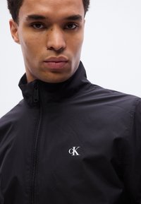 Μαύρο ζακέτα με φερμουάρ από λείο υλικό, με ψηλό γιακά και μικρό λευκό λογότυπο "cK" στην αριστερή πλευρά.