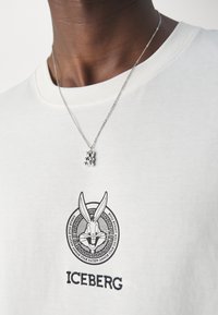 Biała koszulka z okrągłym dekoltem, z czarną grafiką królika i napisem "ICEBERG" poniżej. Srebrny naszyjnik z łańcuszkiem i wisiorkiem.