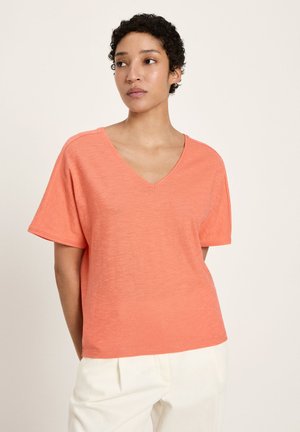 Frau mit kurzen lockigen Haaren, die ein lockeres korallenfarbenes V-Ausschnitt-T-Shirt mit kurzen Ärmeln und weiße Hosen trägt, steht vor einem schlichten hellen Hintergrund.