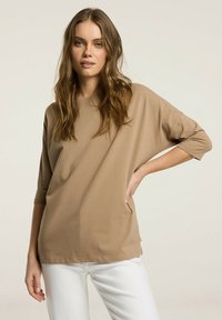 Beżowy, oversize t-shirt z rękawami o długości trzech czwartych, z miękkiej tkaniny i lekko teksturowanym wykończeniem, w połączeniu z białymi spodniami.