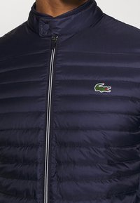 Mörkblå puffjacka med hög krage, horisontellt quiltad textur och dragkedja framtill med en kontrasterande rand. Inkluderar en grön krokodil-logotyp.