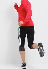 Czerwony top z długim rękawem połączony z czarnymi legginsami capri z białymi paskami. Czarne buty do biegania z teksturowaną podeszwą; dynamiczna poza biegacza.