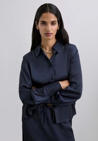 Camisa de botones a rayas azul marino con mangas largas y detalle de cintura atada, que presenta una textura suave y un corte relajado.