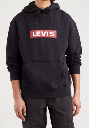 Osoba ubrana w czarną bluzę z kapturem Levi's z czerwono-białym logo, przednią kieszenią i ściągaczami, w połączeniu z czarnymi spodniami.