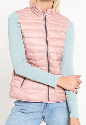 Person wearing a light pink quilted vest over a long-sleeve pale blue shirt and light blue jeans, with one arm crossed over the body.