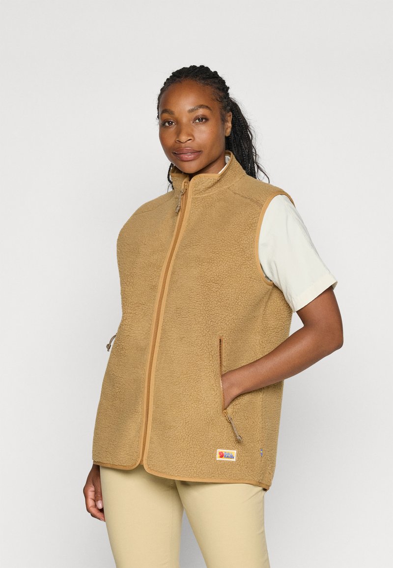 Moteris dėvi rusvos spalvos be rankovių vilnonę liemenę ant baltos trumpomis rankovėmis marškinėlių, viena ranka įkišusi į liemenės kišenę su užtrauktuku.