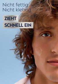 Close-up of a person’s face with light brown, curly hair and a small amount of cream on the cheek against a blue background.