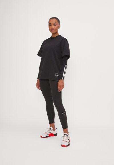 T-shirt noire surdimensionnée avec un logo, associée à un legging noir portant un logo, et des baskets blanches avec des accents rouges et des lacets.