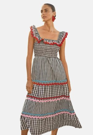 Kvinde iført sort-hvid ternet ærmeløs kjole med røde, pink og blå flæsedetaljer og store røde øreringe.