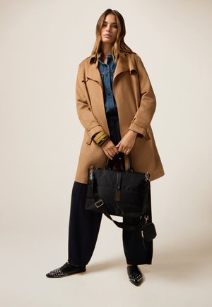 Tan trenchcoat over een denim shirt, gecombineerd met losse marineblauwe broeken en gestifte zwarte flats. Een gewatteerde zwarte tas met verstelbare schouderband wordt vastgehouden.