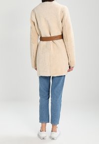 Abrigo beige de pelo sintético con mangas largas, ceñido en la cintura con un cinturón de cuero marrón; combinado con vaqueros azul claro y zapatos blancos.