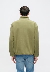 Zelený fleecový pulóver s vysokým golierom a dlhými rukávmi, ktorý má jemnú textúru a voľný strih. Nosí sa cez modré džínsy.