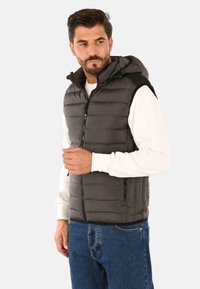 Gilet grigio imbottito da uomo con cappuccio, dotato di una zip frontale, accenti neri sulle spalle e tasche laterali. Indossato sopra una camicia bianca a maniche lunghe.