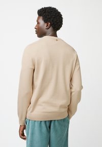 Beige gebreide trui met een ronde halslijn, geribde manchetten en een enkele knoopdetail op de achterkant, gecombineerd met gestructureerde teal broek.