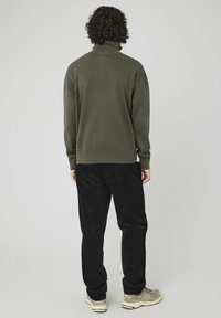 Pull olive avec col haut, texture côtelée et épaules tombantes, associé à un pantalon en velours côtelé noir et des baskets beiges.