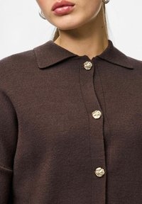 Brązowy dzianinowy kardigan z kołnierzykiem, złotymi kwiatowymi guzikami i fakturowaną tkaniną. Zbliżenie na szczegóły guzika i linię szyi.