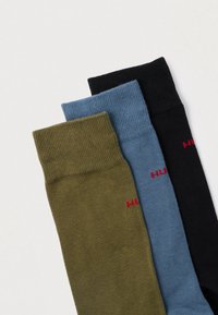 Trois paires de chaussettes : vert olive, bleu clair et noir. Fabriquées en maille texturée, chacune présente un petit logo rouge sur le côté.