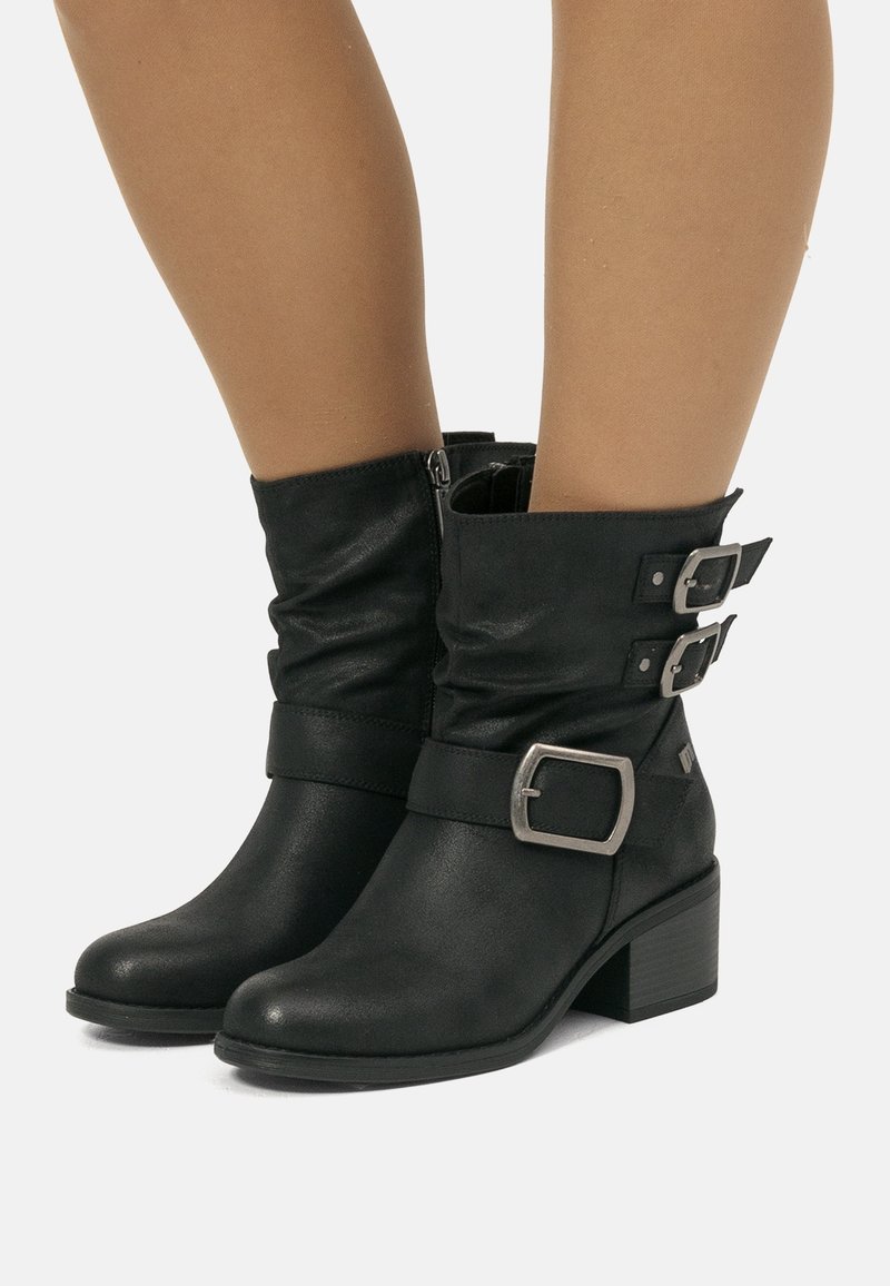 Bottes en cuir noir mi-mollet avec trois boucles argentées et talons carrés, portées sur des jambes nues, sur un fond uni.