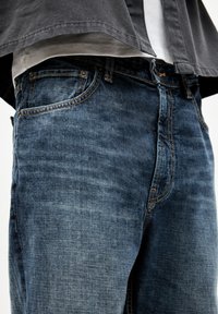 Jeans aus Denim in mittlerem Blau mit einer verblassten Textur. Merkmale sind fünf Taschen, sichtbare Nähte und ein Knopfverschluss an der Taille.