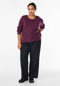 Ciemnofioletowy sweter dzianinowy z okrągłym dekoltem i podwiniętymi rękawami, zestawiony z luźnymi czarnymi dżinsami i czarnym obuwiem.