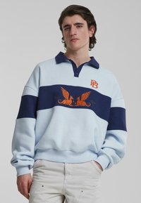 Lyseblå genser med lange ermer, med marineblå striper, krage, brodert logo og design av vinget skapning i oransje. Myk stoff og avslappet passform.
