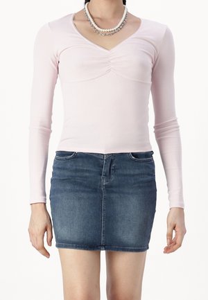 Woman wearing a light pink long-sleeve ruched top, layered pearl and chain necklaces, and a blue denim mini skirt.