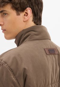 Veste marron avec un col haut, présentant une étiquette en cuir à l'arrière. Texture lisse et design épuré.