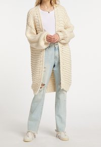 Chunky gebreid crème vest met een lange open voorkant, geribbelde textuur en oversized mouwen, gecombineerd met lichtblauwe distressed jeans en witte sneakers.