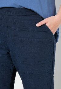 Pantaloni effetto tessuto blu navy con motivo geometrico, dotati di una tasca laterale. Il materiale sembra elastico e morbido al tatto.