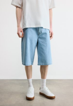 Inimene, kes kannab helerohelisest teksast põlveni ulatuvaid lühikesi pükse, valgeid tavalisi sokke, valgeid tossusid pruunide taldadega ja valget üle mõõdu suuremat T-särki.