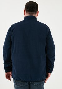 Tamsiai mėlyna flisinė striukė su aukštu apykakle ir ilgomis rankovėmis, pasižyminti lygiu paviršiumi ir laisvu kirpimu, matoma iš nugaros.