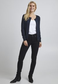 Marinblå cardigan i stickad tyg, med knappframsida och långa ärmar, ihop med svarta smala jeans och ankelboots.