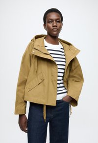 Sárga kapucnis dzseki húzózsinórokkal, oldalzsebekkel és patentos részletekkel, fehér és tengerészkék csíkos pulóver fölött viselve. Sötét farmernadrág látható.