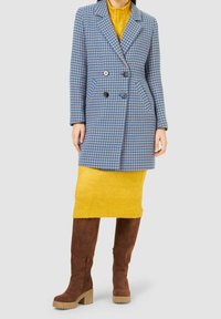 Dvouvrstvý kabát v modrém kostkovaném vzoru s velkými černými knoflíky, doplněný žlutou sukní a hnědými kozačkami ke kolenům.