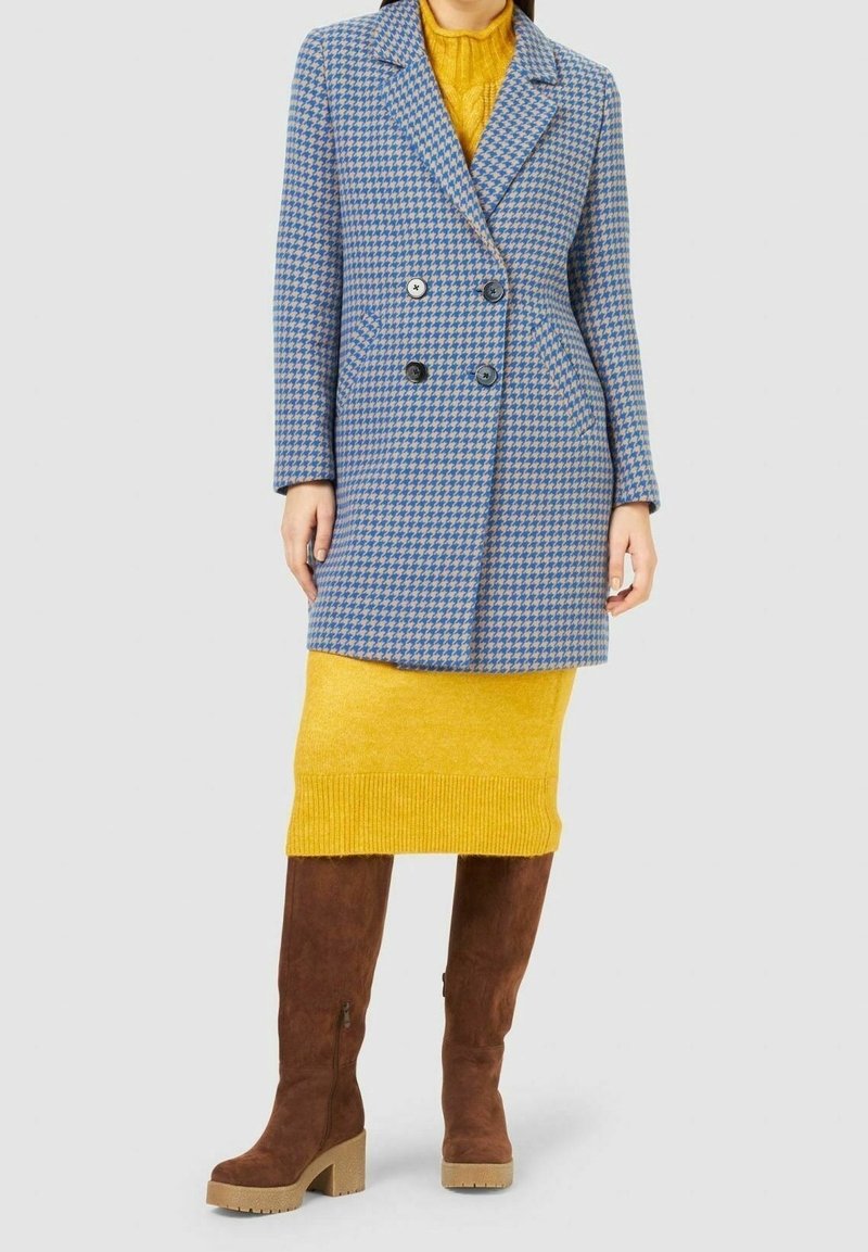 Dvouvrstvý kabát v modrém kostkovaném vzoru s velkými černými knoflíky, doplněný žlutou sukní a hnědými kozačkami ke kolenům.